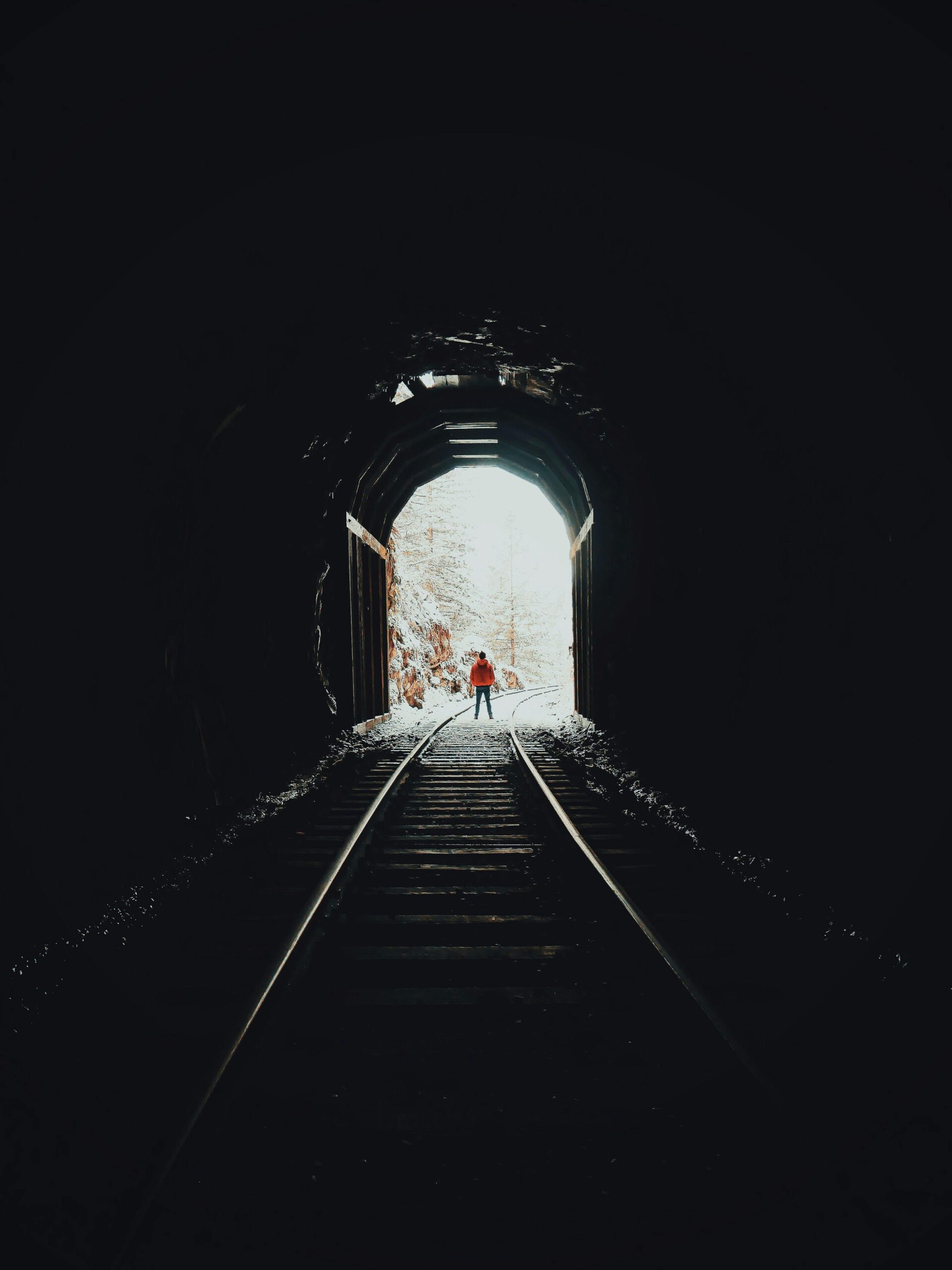 Pessoa de casaco laranja caminhando nos trilhos de trem dentro de um túnel escuro, com saída iluminada por neve branca e árvores ao fundo.