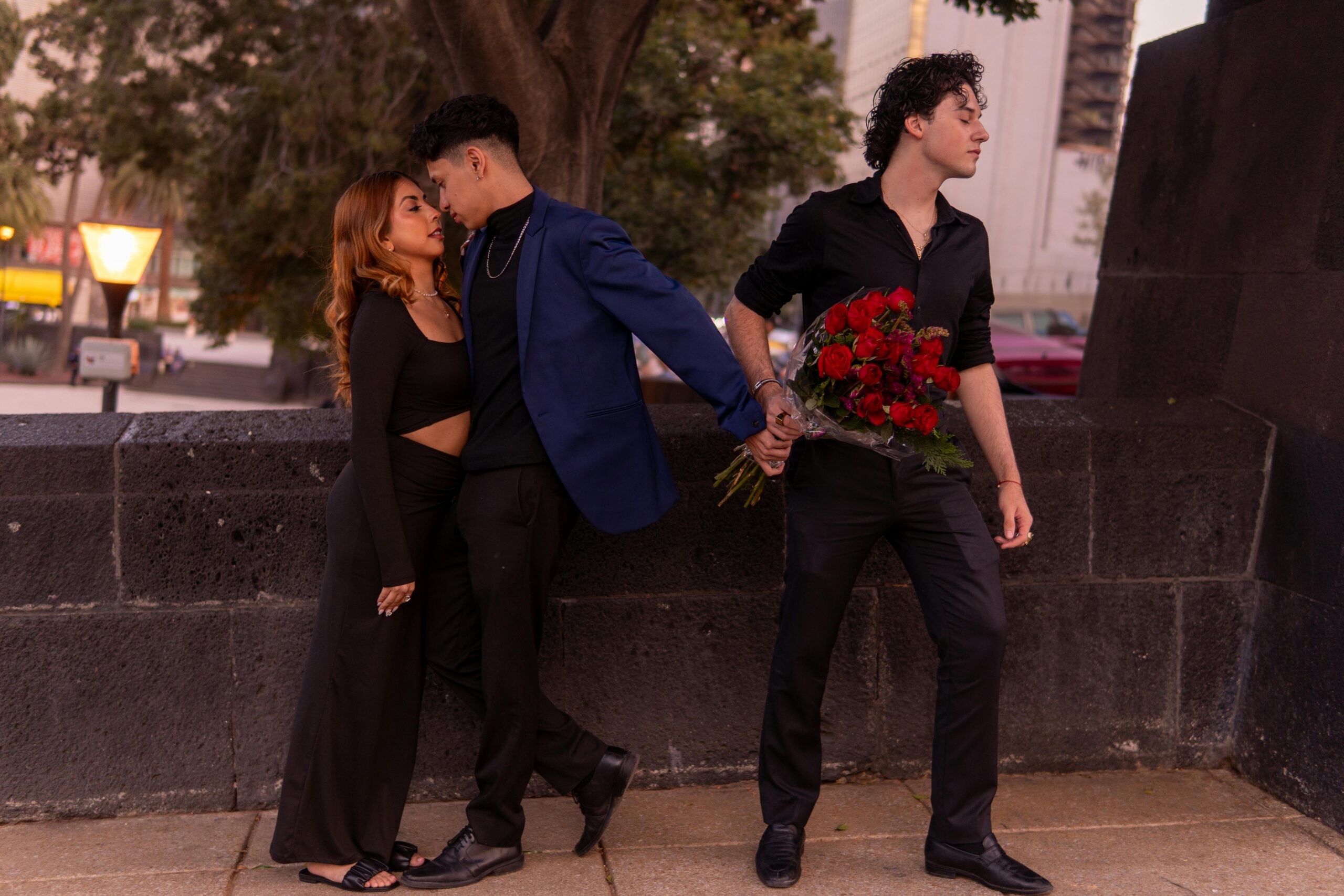 Homem observa casal se beijando enquanto segura buquê de rosas e mantém uma das mãos entrelaçada com o homem do casal, sugerindo um triângulo amoroso e simbolizando traição.