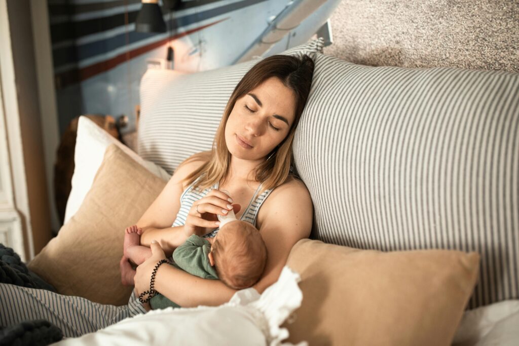 Mãe jovem alimentando bebê recém-nascido no colo enquanto descansa em uma cama, representando cuidado, conexão emocional e início de um novo ciclo.
