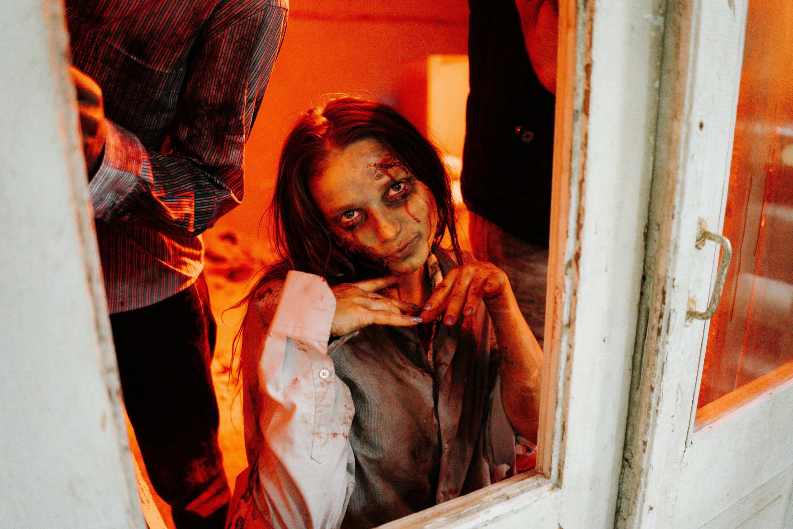 Uma mulher transformada em zumbi, com olhos esbranquiçados e expressão sinistra, está dentro de uma casa, vista por uma janela branca. Ela posa de forma irônica, com as mãos sob o queixo, como se estivesse zombando. Sua pele está pálida e suja, com manchas de sangue no rosto e nas roupas. O ambiente está iluminado por uma forte luz alaranjada, sugerindo perigo iminente ou incêndio. Ao redor dela, outros zumbis são parcialmente visíveis.