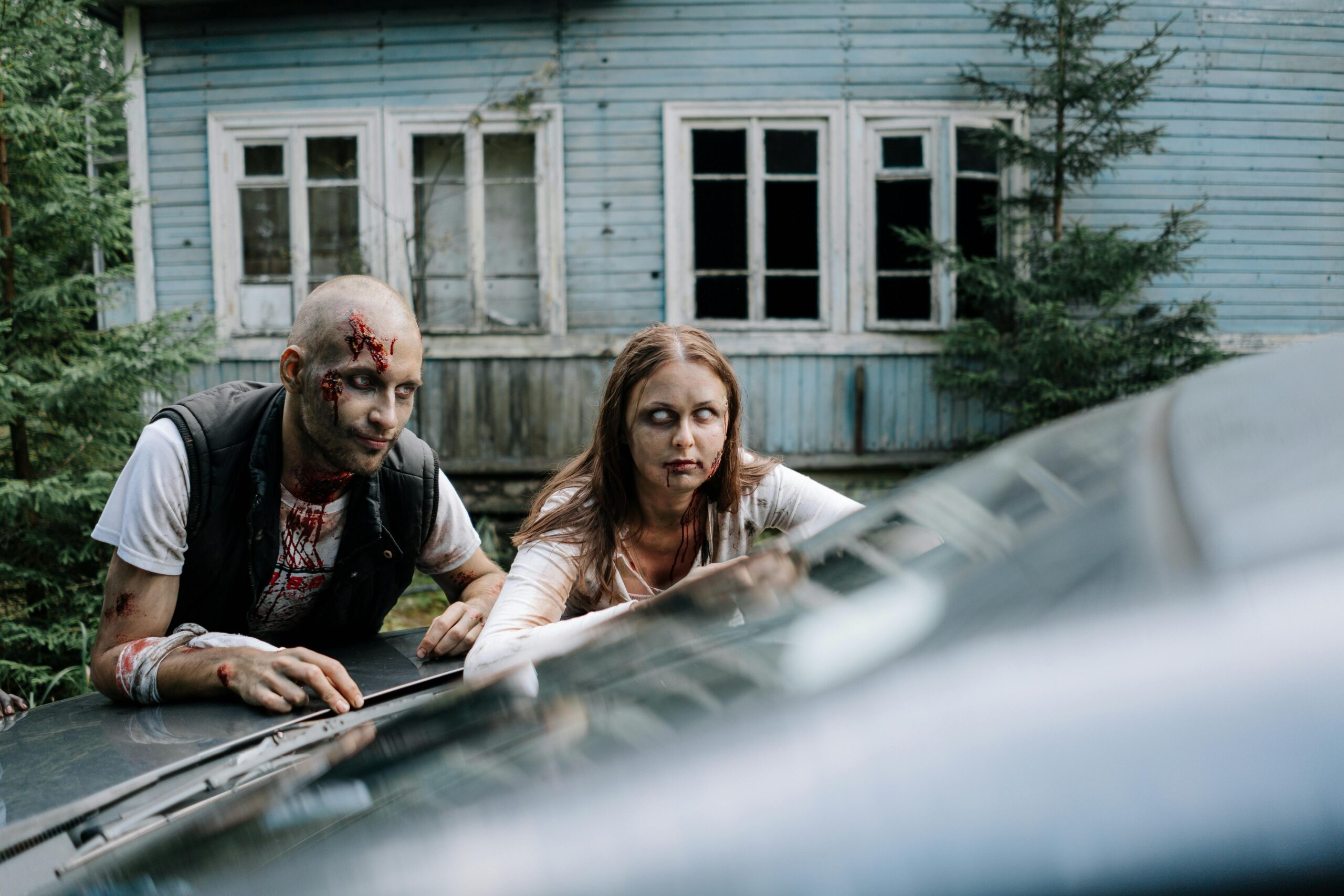 Dois zumbis estão próximos a um carro. O zumbi à esquerda é um homem careca com cortes profundos e sangue escorrendo no rosto, vestindo camiseta branca manchada de sangue e colete preto. À direita, uma mulher com olhos brancos e expressão vazia está apoiada no capô do carro. Ambos estão em frente a uma casa de madeira azul em ruínas, com janelas quebradas e cercados por vegetação. A cena transmite abandono e terror.