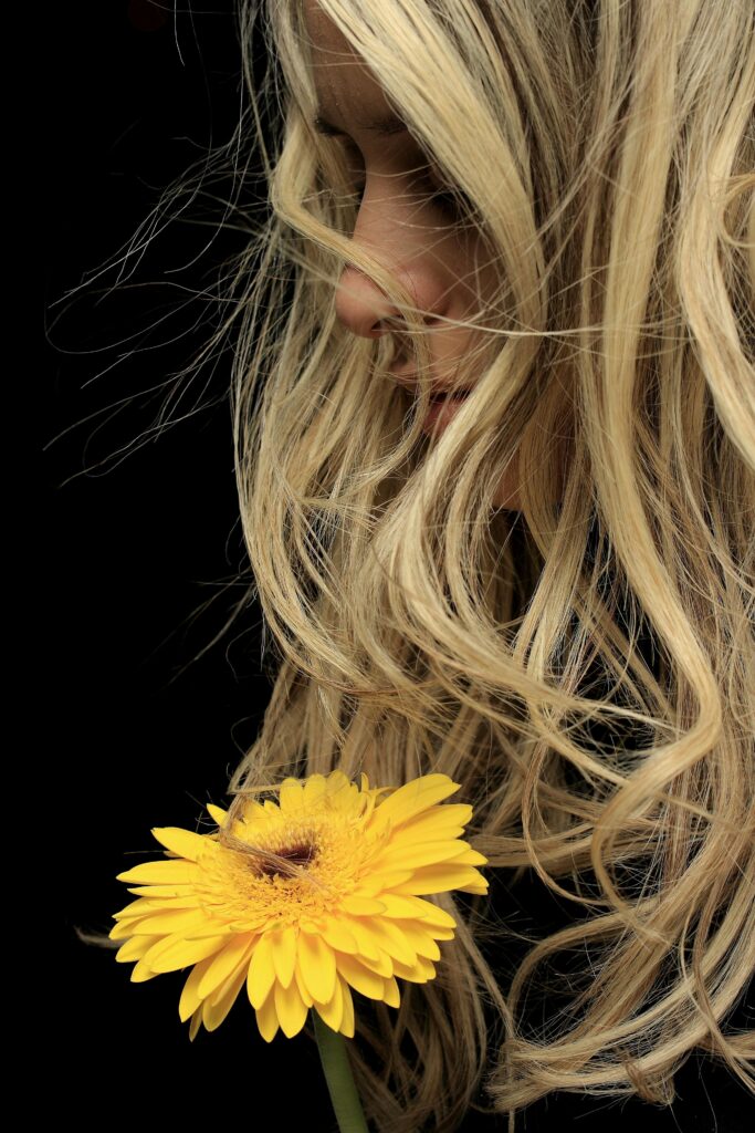 Mulher loira de cabelos longos e ondulados próxima a uma flor amarela, representando beleza e energia positiva nos sonhos.