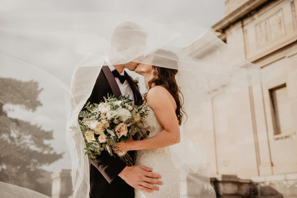 Casal de noivos se beijando sob o véu, com a noiva segurando um buquê de flores e o véu suavemente esvoaçando ao vento.