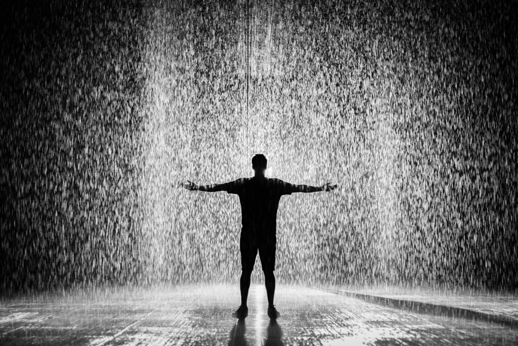 Pessoa em pé com braços abertos sob forte chuva, em um ambiente escuro, representando sonhar com chuva intensa.