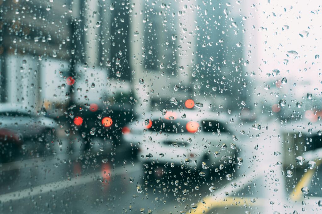 Vidro de carro com gotas de chuva, mostrando uma rua movimentada ao fundo, relacionado a sonhar com chuva.